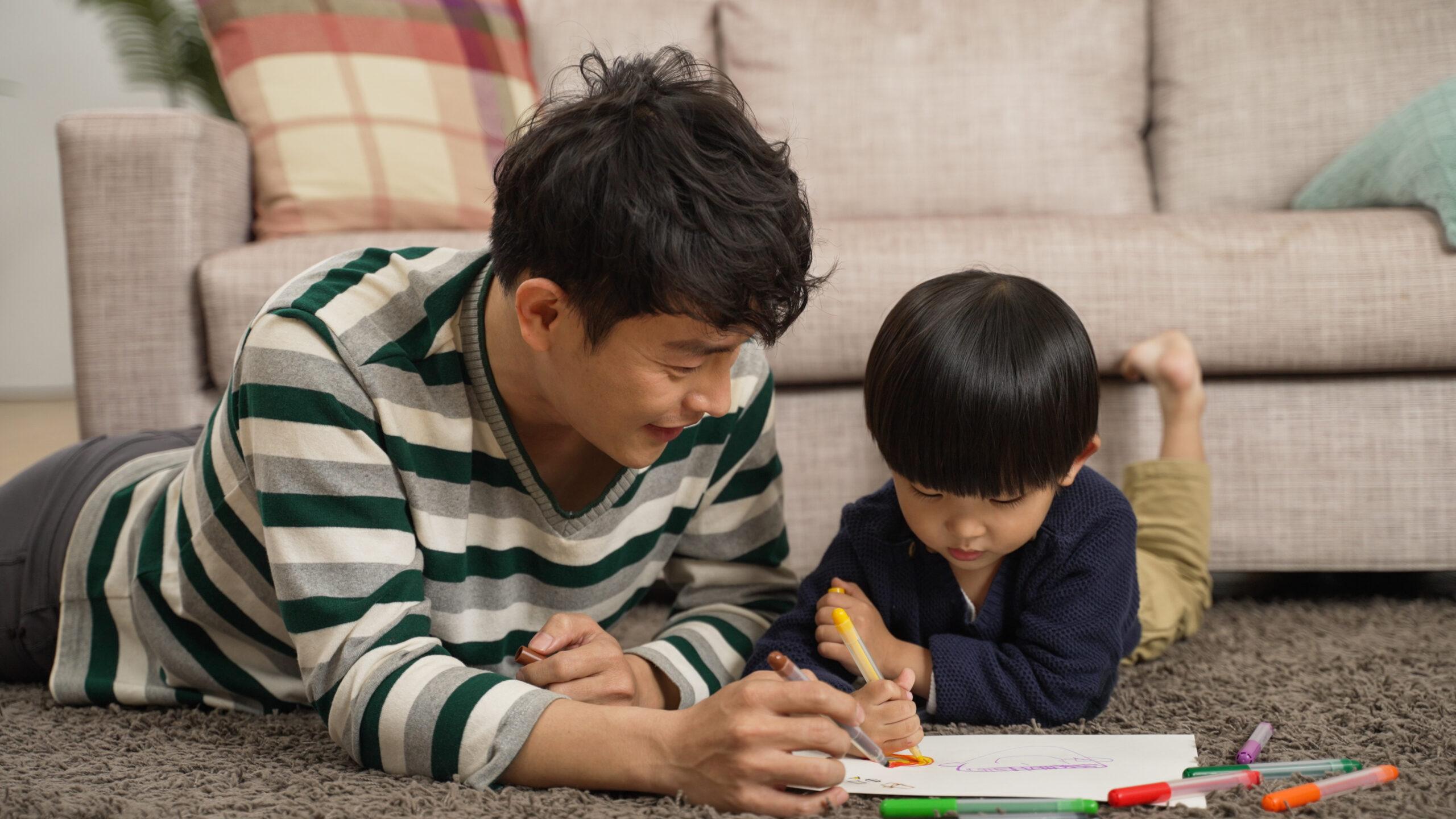 loving father writing with his baby son while lying on belly on floor